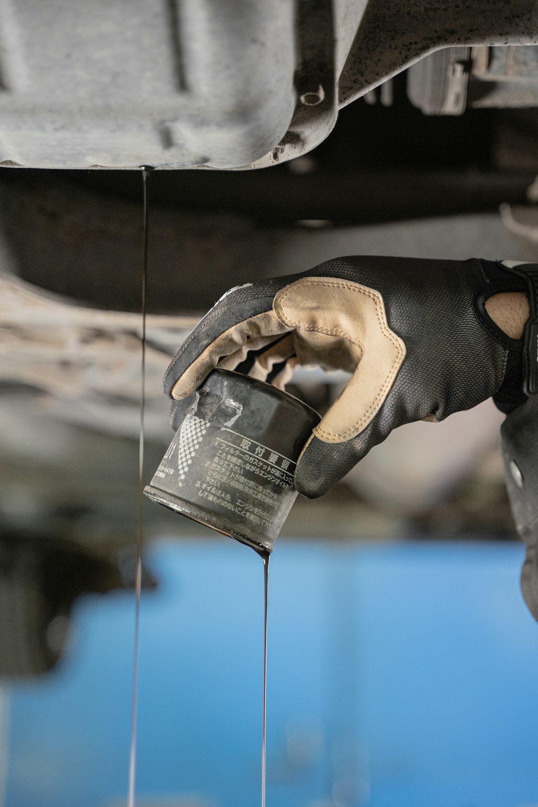 Gloved hand removing an oil filter under a vehicle while oil runs down in a thin line